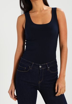 Mujer con camiseta sin mangas azul marino ajustada combinada con jeans de talle alto azul oscuro sobre un fondo liso.