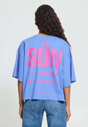 Frau mit langen lockigen dunklen Haaren, die ein lockeres hellblaues Hemd mit pinkem Text „THE SUN IS MY MUSE“ und blaue Jeans trägt, blickt nach hinten.