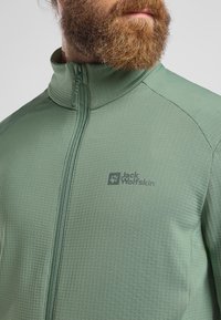 Veste zippée verte avec une finition texturée, logo sur le côté gauche, col montant et fermeture éclair contrastante, conçue pour les activités de plein air.