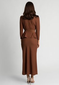 Długa brązowa sukienka z peplum w talii, długimi rękawami i dopasowanym krojem. Gładka tekstura, w połączeniu z jasnymi obcasami.