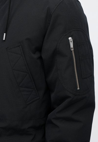 Artimiausias juodo švarko rankovės vaizdas su sidabriniu užtrauktuku kišenėje ir siuvinėtu stačiakampiu pleistru ant rankos.