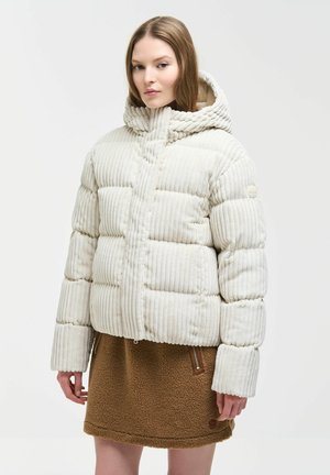 Frau trägt eine weiße, gerippte Steppjacke mit hohem Kragen und Kapuze, kombiniert mit einem braunen, strukturierten Rock mit Reißverschlusstaschen.