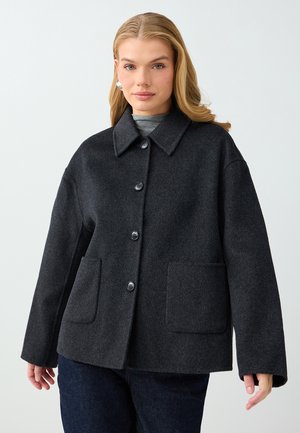 Une femme présente un manteau gris anthracite foncé, boutonné sur le devant, avec un large col et deux poches plaquées, sur un fond clair uni.
