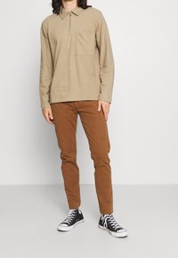 Chemise beige à manches longues avec col, fermeture éclair et poche poitrine, associée à un pantalon marron ajusté et des baskets noires basses.