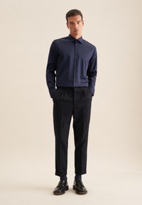 Camicia blu navy con tessuto morbido, abbinata a pantaloni neri plissettati. Le scarpe sono in pelle nera con finitura lucida.
