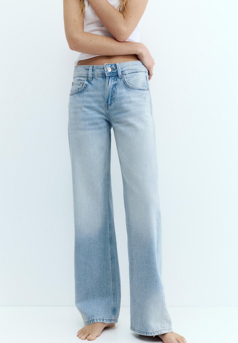 Žena u svijetloplavim trapericama visokog struka širokih nogavica i bijeloj bezrukavnoj majici, stoji bosonoga na bijeloj pozadini.