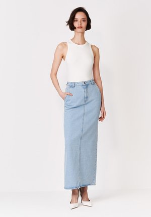 Žena s krátkymi tmavými vlasmi, oblečená v bielom tielku bez rukávov, svetlomodrej dlhé rifľovej sukni s vysokým pásom a bielych špicatých lodičkách, stojaca pred bielym pozadím.