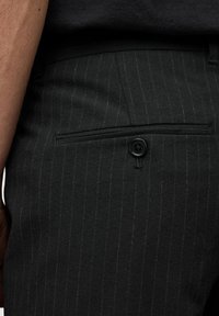 Czarne spodnie w prążki z tylną kieszenią, z zapięciem na czarny guzik i gładką teksturą. Widoczna dostosowana talia.