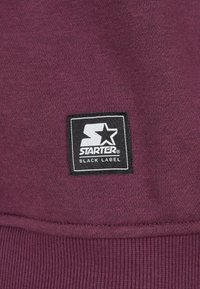 Tela morada con un parche del logo Starter Black Label en blanco y negro cosido cerca del borde inferior de la prenda.