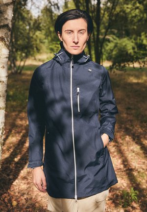 Navy waterproof jacket with a high collar, front zip closure, and pockets. Features adjustable hardware and a relaxed fit. Forest background.