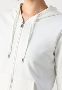 Gros plan d'une personne portant un sweat à capuche blanc zippé avec cordons et poche avant sur un fond uni.