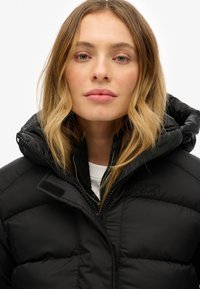 Vrouw met golvend blond haar die een zwarte Superdry gewatteerde jas met capuchon draagt, tegen een effen witte achtergrond.
