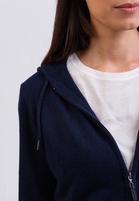 Mörkblå zip-up hoodie med justerbara dragsnören, mjuk textur och en vit t-shirt med rund hals under. Närbild av halslinningen.