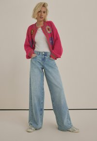 Cardigan lavorato a maglia di un vivace rosa con disegni floreali multicolori, abbinato a jeans a gamba larga azzurri e sneakers argentate.