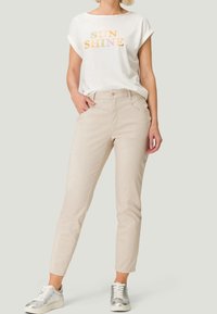 T-shirt blanc à manches courtes avec le texte "SUN SHINE" en orange, associé à un pantalon beige slim-fit et des baskets argentées. Tenue simple et décontractée.