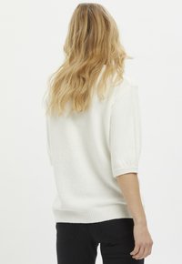 Maglione lavorato a maglia bianco con una vestibilità rilassata, maniche a sbuffo corte, bordo a costine e superficie testurizzata. Indossato con jeans neri, mostrando una vista dal retro.