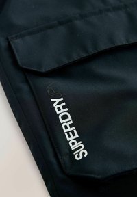 Svart tygficka med en slät yta, med texten "SUPERDRY" i vitt. Fickan har en klaffdesign och en invändig söm.