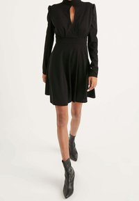 Robe noir avec un col haut, des manches longues et une découpe en forme de clé au niveau de la poitrine. Jupe évasée et tissu texturé. Associée à des bottines noires à la cheville.