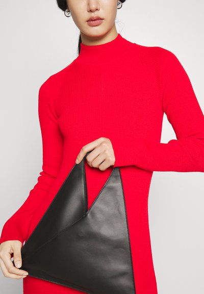 Sac en cuir noir en forme de triangle tenu par une personne portant une robe en turtleneck côtelée rouge, mettant en valeur la texture et le design géométrique.