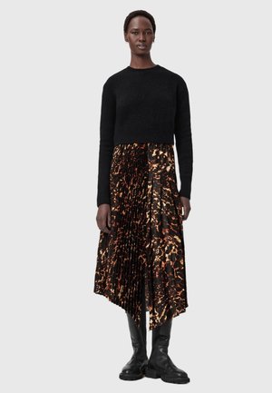 Zwart cropped sweater met lange mouwen, gecombineerd met een geplooide rok met een bruin en oranje abstract patroon. Het model draagt zwarte kniehoge laarzen.