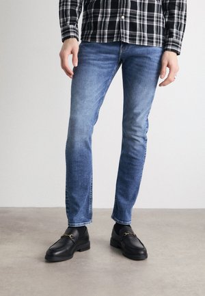 Jeans in denim blu con una vestibilità slim, caratterizzati da un lavaggio sbiadito, leggere sfumature e cuciture a contrasto. Abbinati a mocassini neri.
