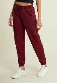 Persona che indossa pantaloni jogger bordeaux con tasche laterali e la scritta "IL MOVIMENTO È UNA TERAPIA", abbinati a scarpe da ginnastica bianche su sfondo beige.