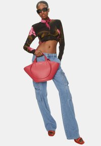 Rode leren handtas met een ronde vorm en dubbele handvatten. Model draagt een multicolored crop top en wijde denim broek, accessorized met een zonnebril.