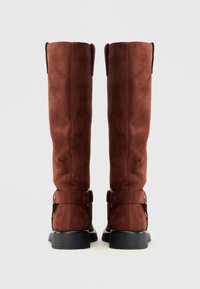 Botas altas de ante marrón con una textura suave, acentos de hebilla ajustable y una suela negra resistente. La vista trasera muestra líneas y costuras limpias.