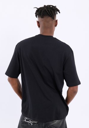 Man met korte dreadlocks, gekleed in een effen zwart T-shirt en gescheurde donkere jeans, staand met handen achter de rug, met de rug naar de camera.