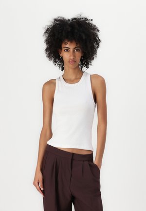 Débardeur blanc à côtes avec un col rond, coupe ajustée et sans manches, associé à un pantalon brun taille haute.