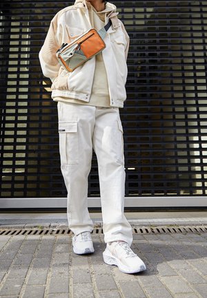 Pantalon cargo blanc avec poches latérales, une veste beige à fermeture éclair et un sac bandoulière vert olive et orange. Baskets blanches avec semelles tachetées.