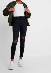 Jeans neri attillati con dettagli strappati sulle ginocchia, abbinati a un top bianco aderente e a una giacca bomber verde oliva. Sneakers bianche completano il look.