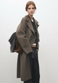 Lang bruin wollen jas met grote revers, gecombineerd met een grijze top en zwarte broek. Bruine suède tas over de schouder gedragen.