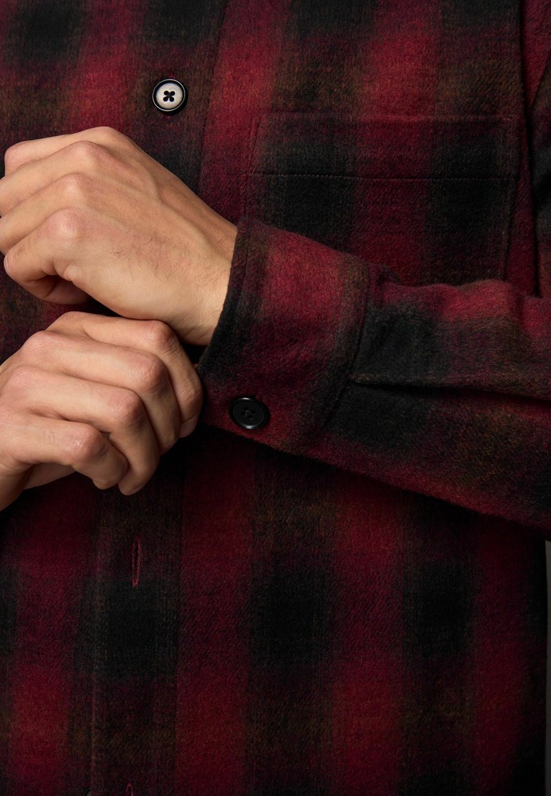 Chemise en flanelle à carreaux rouge et noir avec fermeture à boutons, dotée d'une poche poitrine et d'un tissu doux et texturé. Les boutons sont noirs et ronds.