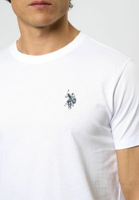 Vit bomullströja med rund halsringning som har en liten blå grafisk bild av två polospelare på hästar. Mjuk textur, klassisk passform.