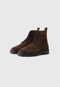 Botas de ante marrón hasta el tobillo con punta redonda, cordones en la parte delantera, perforaciones decorativas de brogue y una suela de goma negra. Presentan lengüetas en la parte superior.