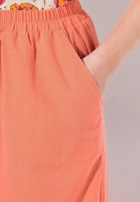 Perzikkleurige stoffen broek met een elastische tailleband en zijzakken. De textuur is glad met een lichte zachtheid. Past nauwsluitend om het lichaam.