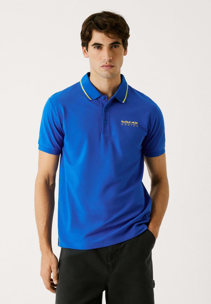 Niebieska koszulka polo z krótkim rękawem, kontrastowym żółtym kołnierzykiem oraz haftowanym logo Red Bull Racing na piersi. Gładka faktura materiału.
