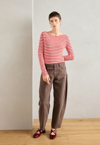 Červeno-biele pruhované tričko s dlhým rukávom a lodičkovým výstrihom, v kombinácii s voľnými hnedými nohavicami a červenými topánkami s pásikmi.