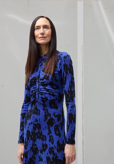 Robe longue à manches longues bleue avec imprimé abstrait noir, présentant des détails froncés sur le devant et une silhouette ajustée. Texture de tissu lisse.