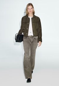 Giacca in suede verde oliva con un design aderente, abbinata a jeans a zampa d'elefante grigi chiari. Gli accessori includono una borsa nera e una semplice collana con pendente.