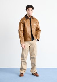 Chaqueta de cuero marrón con cierre delantero, cuello oscuro en contraste y bolsillos laterales, llevada sobre una camisa negra y pantalones beiges.