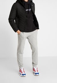Un giubbotto trapuntato nero, una felpa con cappuccio nera con logo, pantaloni della tuta grigi e scarpe da ginnastica rosse, bianche e blu creano un look casual stratificato.