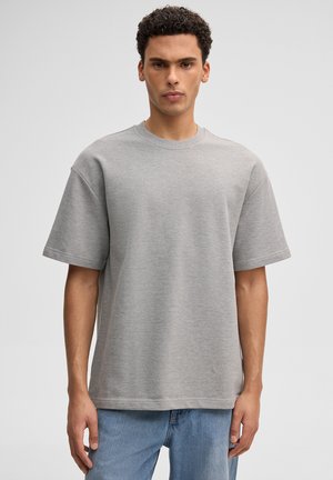 Jonge man met kort krullend haar, gekleed in een effen lichtgrijs oversized T-shirt met korte mouwen en een blauwe spijkerbroek, staand tegen een lichte achtergrond.