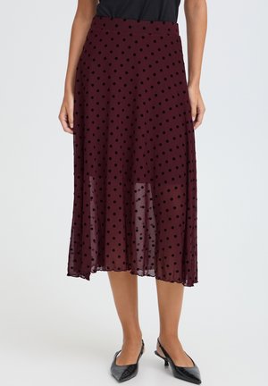 Femme portant une jupe midi bordeaux à pois noirs et des chaussures slingback noires pointues à talons bas, debout, les mains le long du corps.