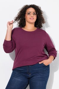 Frau mit lockigem Haar, die eine Brille trägt, ein lila Langarmshirt und blaue Jeans anhat, hält eine Haarsträhne mit einer Hand und hat die andere in der Tasche.