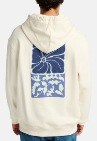 Haut à capuche cream avec un grand graphique floral et de feuilles bleu au dos, avec des détails texturés et un ourlet côtelé.