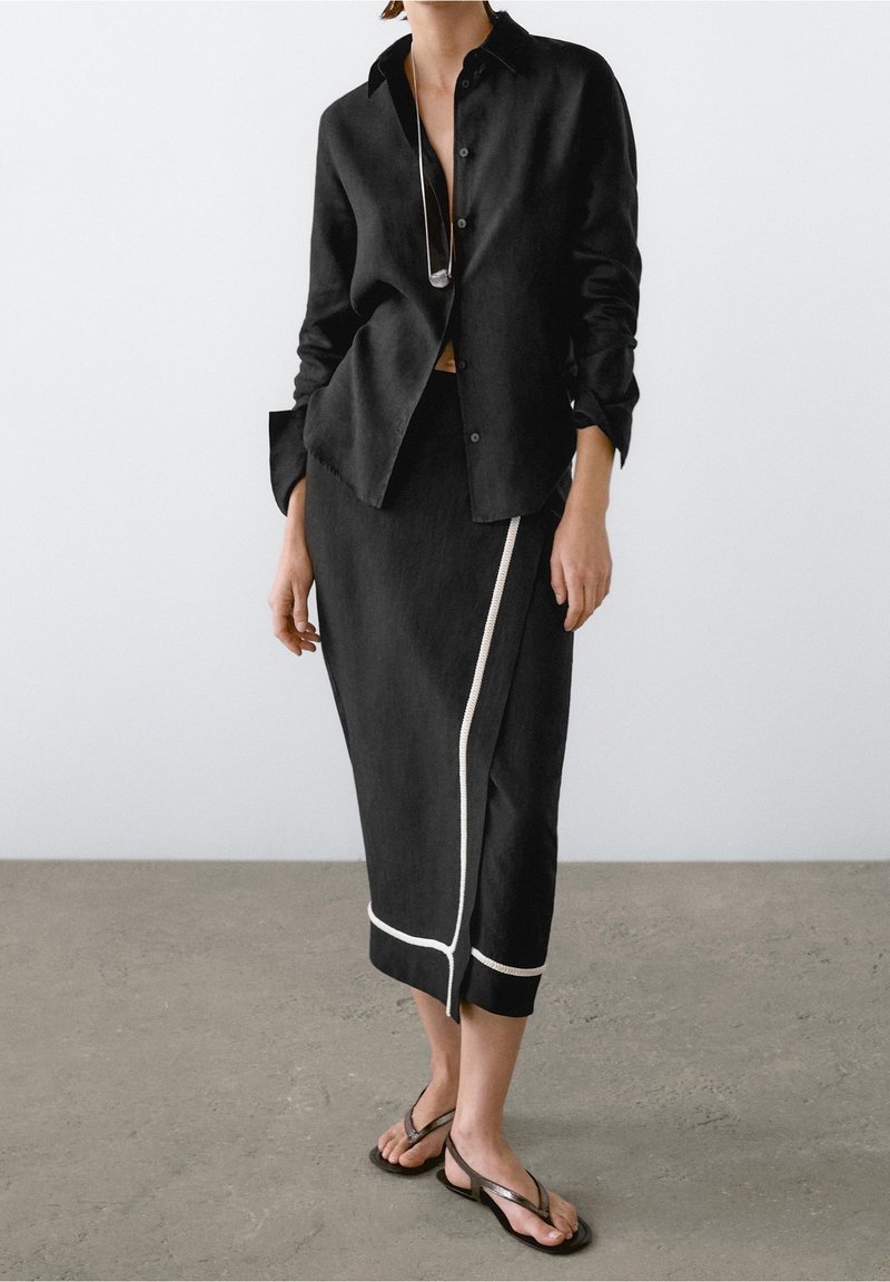 Femme portant une chemise noire boutonnée, une jupe midi noire avec un liseré blanc et des sandales noires, debout contre un mur gris clair uni.