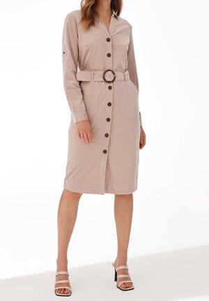 Femme portant une robe chemise beige ceinturée avec des boutons sur le devant, manches longues et sandales à talons beige, debout devant un fond blanc.