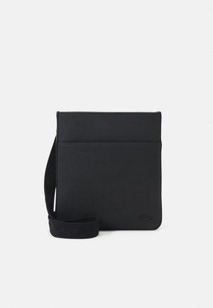Sac bandoulière rectangulaire noir avec une large sangle et un logo embossé subtil dans le coin inférieur sur un fond clair uni.